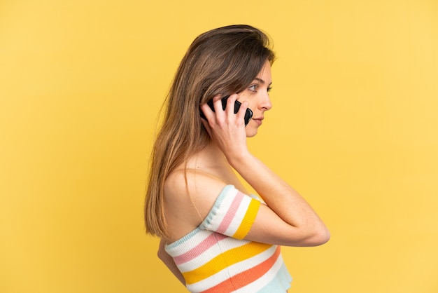 Jeune femme caucasienne isolée sur fond jaune gardant une conversation avec le téléphone portable avec quelqu'un