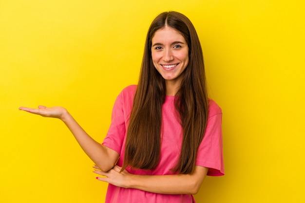 Jeune femme caucasienne isolée sur fond jaune montrant un espace de copie sur une paume et tenant une autre main sur la taille.