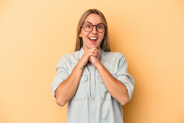 Jeune femme caucasienne isolée sur fond jaune priant pour la chance, étonnée et ouvrant la bouche regardant vers l'avant.