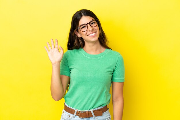 Jeune femme caucasienne isolée sur fond jaune saluant avec la main avec une expression heureuse