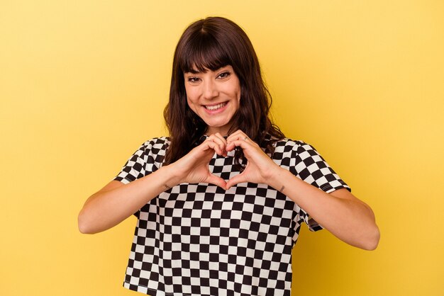 Jeune femme caucasienne isolée sur fond jaune souriant et montrant une forme de coeur avec les mains.