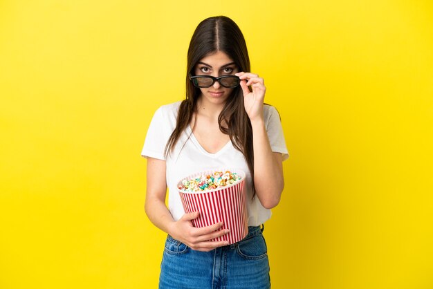 Jeune femme caucasienne isolée sur fond jaune surprise avec des lunettes 3d et tenant un grand seau de pop-corn