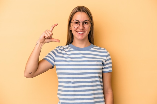 Jeune femme caucasienne isolée sur fond jaune tenant quelque chose de petit avec les index, souriante et confiante.