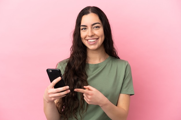 Jeune femme caucasienne isolée sur fond rose à l'aide de téléphone mobile et en le pointant