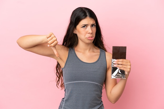 Jeune femme caucasienne isolée sur fond rose prenant une tablette de chocolat faisant un mauvais signal