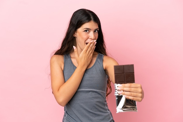 Jeune femme caucasienne isolée sur fond rose prenant une tablette de chocolat et surprise