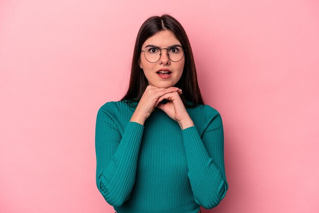 Jeune femme caucasienne isolée sur fond rose priant pour la chance étonnée et ouvrant la bouche en regardant vers l'avant