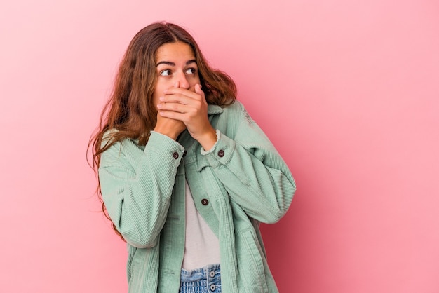 Jeune femme caucasienne isolée sur fond rose réfléchie à la recherche d'un espace de copie couvrant la bouche avec la main.