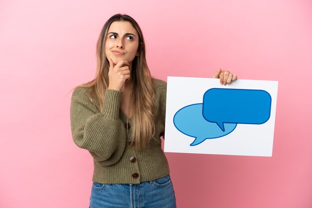 Jeune femme caucasienne isolée sur fond rose tenant une pancarte avec l'icône de la bulle de dialogue et la pensée