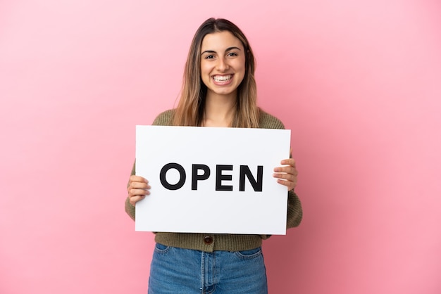 Jeune femme caucasienne isolée sur fond rose tenant une pancarte avec texte OUVERT avec une expression heureuse