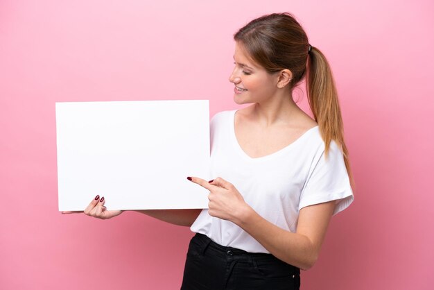 Jeune femme caucasienne isolée sur fond rose tenant une pancarte vide avec une expression heureuse et la pointant