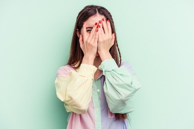 Jeune femme caucasienne isolée sur fond vert clignote devant la caméra à travers les doigts, embarrassée couvrant le visage.