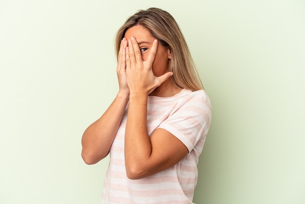 Jeune femme caucasienne isolée sur fond vert clignote à travers les doigts effrayés et nerveux.