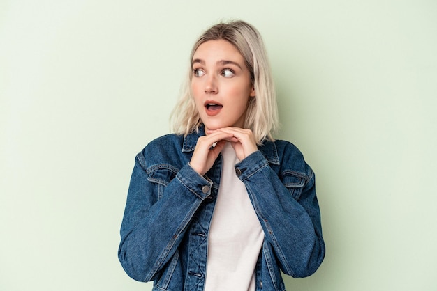 Jeune femme caucasienne isolée sur fond vert priant pour la chance, étonnée et ouvrant la bouche regardant vers l'avant.