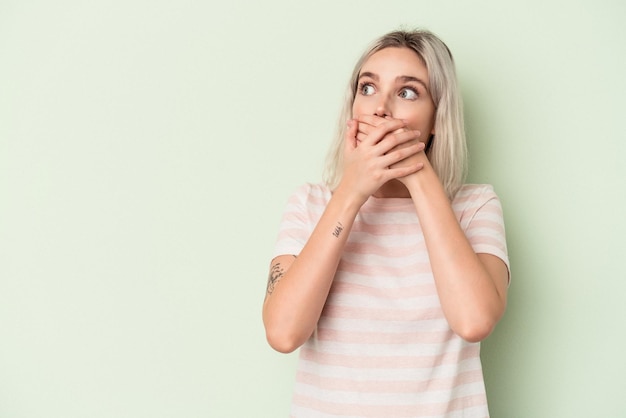 Jeune femme caucasienne isolée sur fond vert réfléchie à la recherche d'un espace de copie couvrant la bouche avec la main.