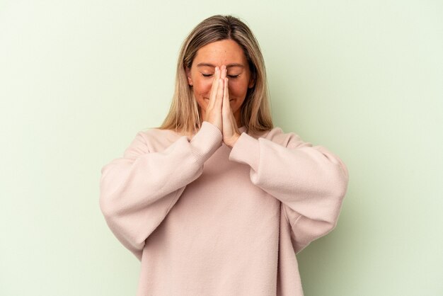 Jeune femme caucasienne isolée sur fond vert tenant la main en prière près de la bouche, se sent confiante.