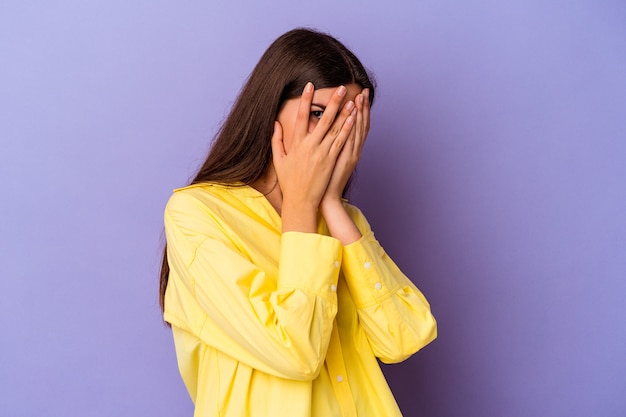 Jeune femme caucasienne isolée sur fond violet clignote à travers les doigts effrayés et nerveux.