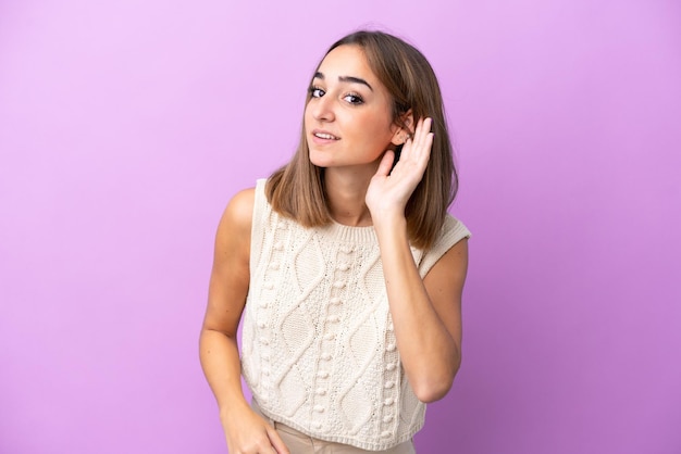 Jeune femme caucasienne isolée sur fond violet écoutant quelque chose en mettant la main sur l'oreille