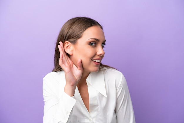Jeune femme caucasienne isolée sur fond violet écoutant quelque chose en mettant la main sur l'oreille