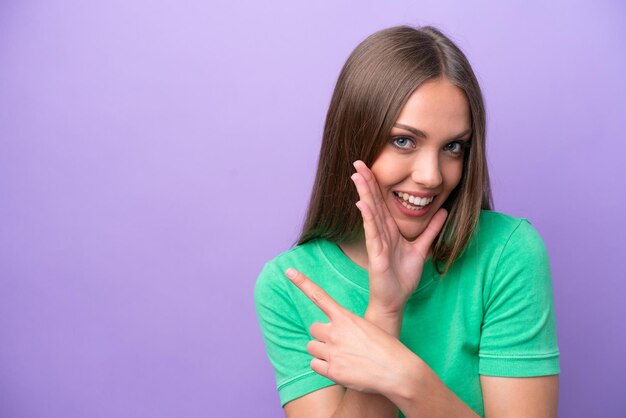 Jeune femme caucasienne isolée sur fond violet pointant vers le côté pour présenter un produit et chuchotant quelque chose