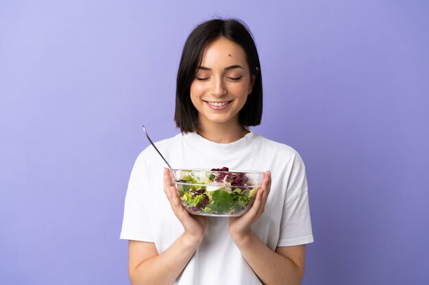 Jeune femme caucasienne isolée sur fond violet tenant un bol de salade avec une expression heureuse