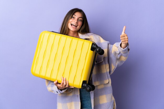 Jeune femme caucasienne isolée sur fond violet en vacances avec valise de voyage