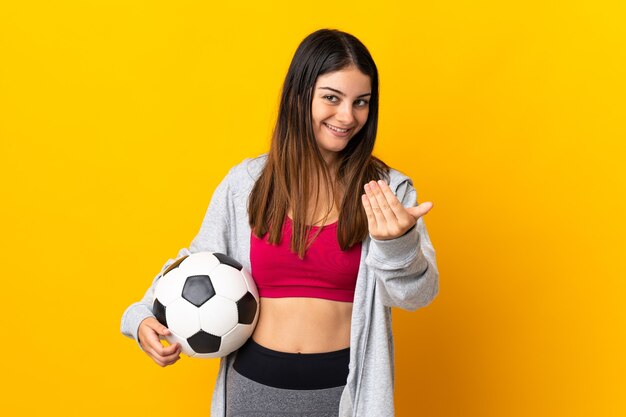 Jeune femme caucasienne isolée sur jaune avec ballon de foot et faire le geste à venir