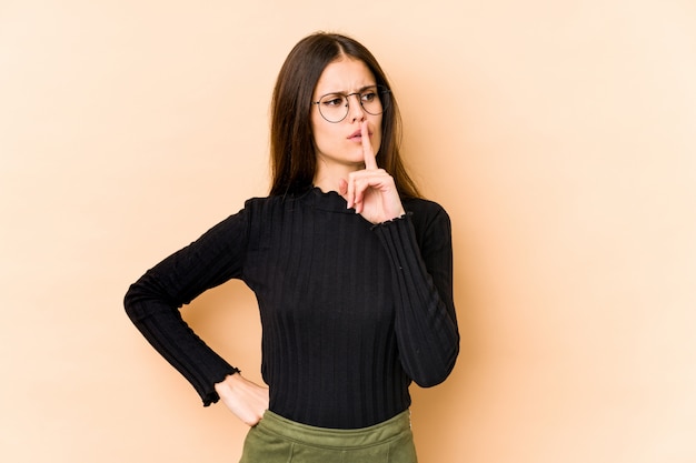 Jeune femme caucasienne isolée sur mur beige gardant un secret ou demandant le silence.