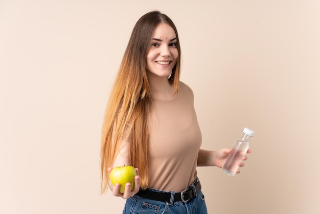 Jeune femme caucasienne isolée sur mur beige avec une pomme et avec une bouteille d'eau