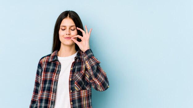 Jeune femme caucasienne isolée sur mur bleu avec les doigts sur les lèvres gardant un secret.