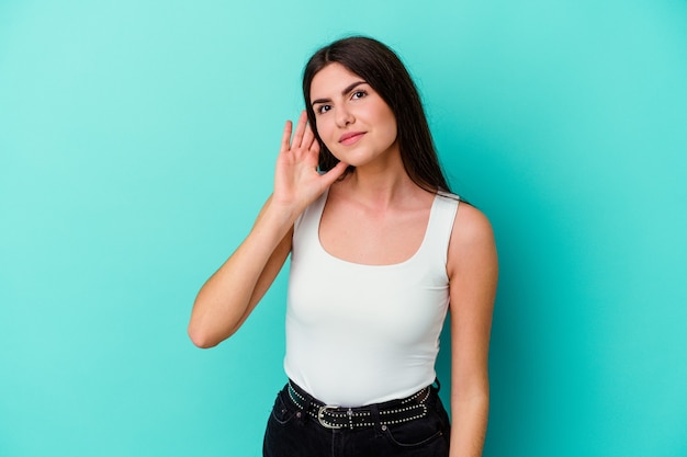 Jeune femme caucasienne isolée sur un mur bleu essayant d'écouter un potin.