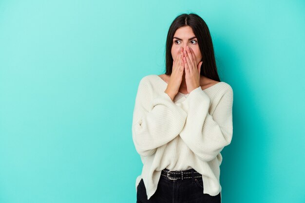 Jeune femme caucasienne isolée sur un mur bleu réfléchie à la recherche d'un espace de copie couvrant la bouche avec la main.