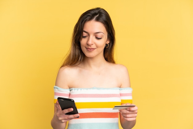 Jeune femme caucasienne isolée sur un mur jaune achetant avec le mobile avec une carte de crédit