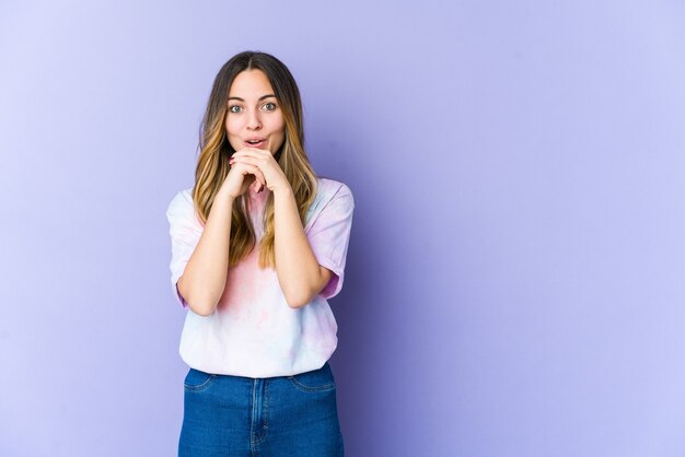 Jeune femme caucasienne isolée sur un mur violet priant pour la chance, étonné et ouvrant la bouche à l'avant.