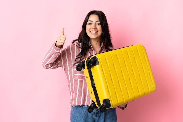 Jeune femme caucasienne isolée sur rose en vacances avec valise de voyage et avec le pouce vers le haut