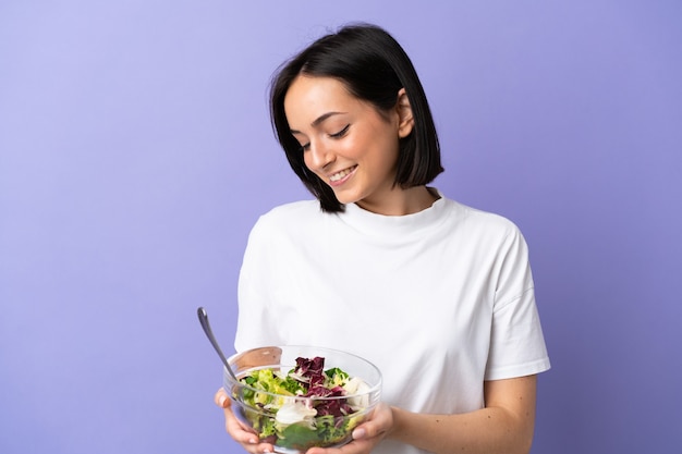 Jeune femme caucasienne isolée sur violet tenant un bol de salade avec une expression heureuse