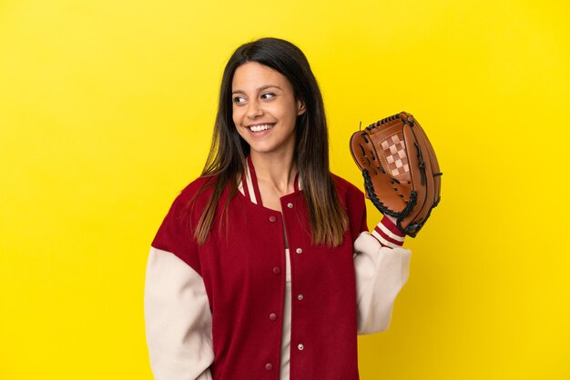 Jeune femme caucasienne jouant au baseball isolé sur fond jaune regardant sur le côté et souriant