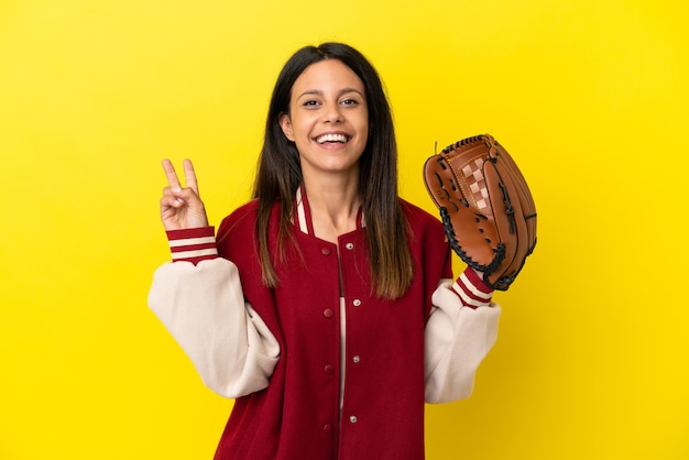 Jeune femme caucasienne jouant au baseball isolé sur fond jaune souriant et montrant le signe de la victoire
