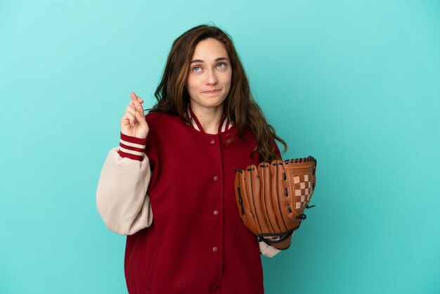 Jeune femme caucasienne jouant au baseball isolée sur fond bleu avec les doigts croisés et souhaitant le meilleur