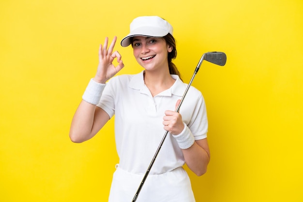 Jeune femme caucasienne jouant au golf isolée sur fond jaune montrant un signe ok avec les doigts