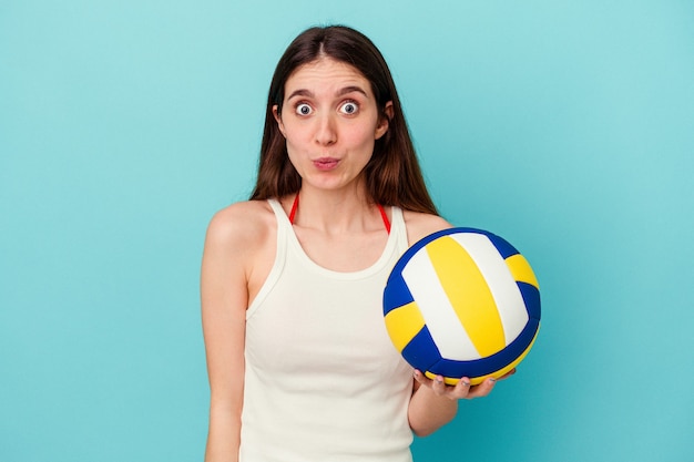 Jeune femme caucasienne jouant au volley-ball isolée sur fond bleu hausse les épaules et ouvre les yeux confus.