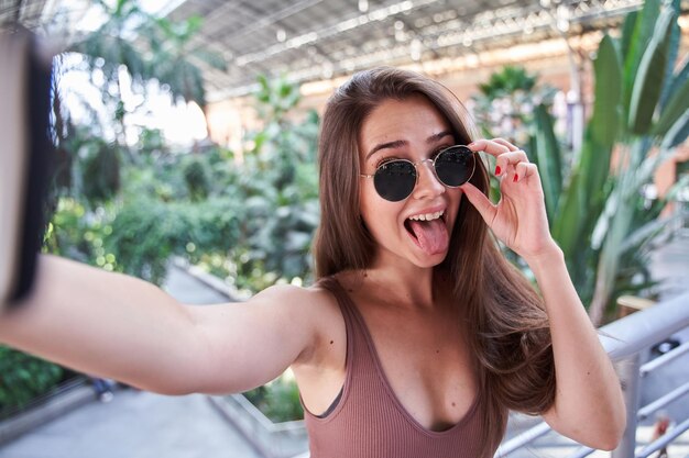 Jeune femme caucasienne avec des lunettes de soleil et des cheveux longs prenant un selfie avec un téléphone intelligent tout en tirant la langue