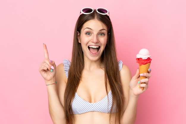 Jeune femme caucasienne en maillot de bain tenant une glace au cornet isolée sur fond rose pointant vers une excellente idée