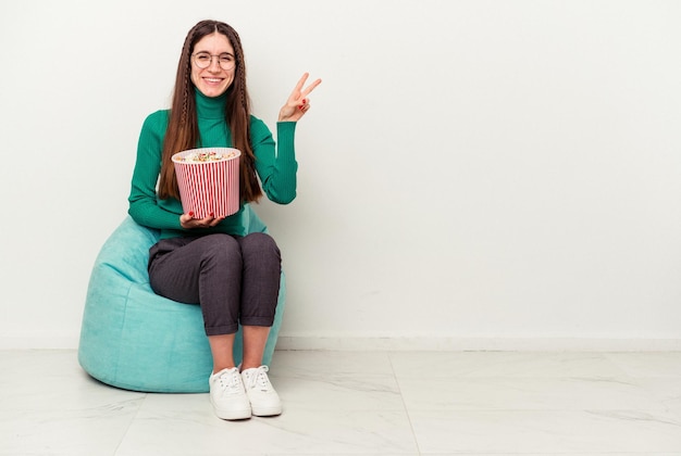 Jeune femme caucasienne mangeant des pop-corns sur une bouffée isolée sur fond blanc joyeuse et insouciante montrant un symbole de paix avec les doigts.