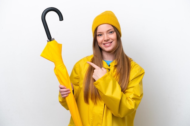 Jeune femme caucasienne avec manteau imperméable et parapluie isolé sur fond blanc pointant vers le côté pour présenter un produit