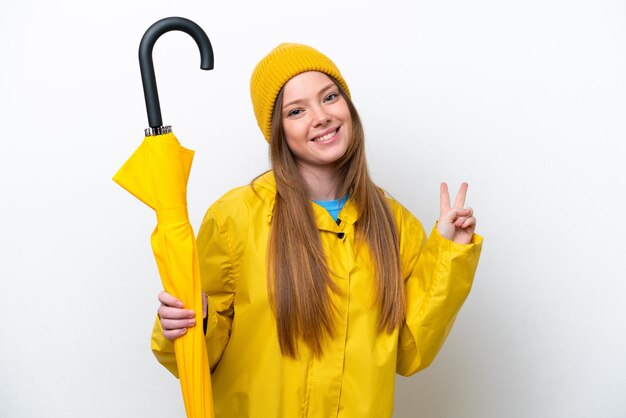 Jeune femme caucasienne avec manteau imperméable et parapluie isolé sur fond blanc souriant et montrant le signe de la victoire