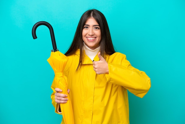 Jeune femme caucasienne avec manteau imperméable et parapluie isolé sur fond bleu donnant un geste du pouce levé