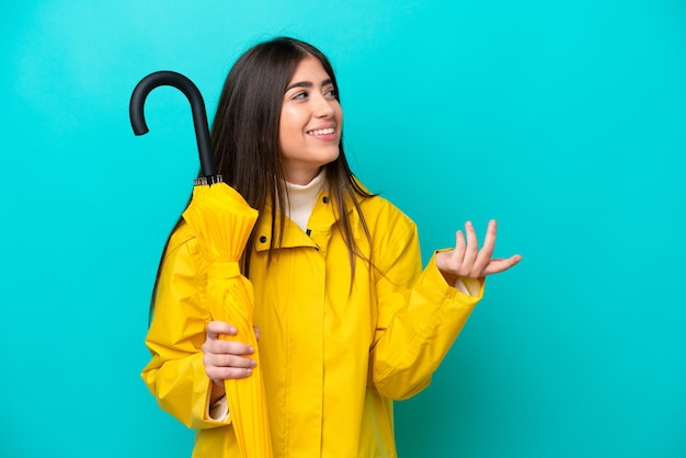 Jeune femme caucasienne avec manteau imperméable et parapluie isolé sur fond bleu tendant les mains sur le côté pour inviter à venir