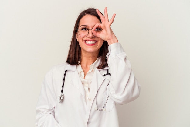 Jeune femme caucasienne médecin isolée sur fond blanc excité en gardant le geste ok sur les yeux.