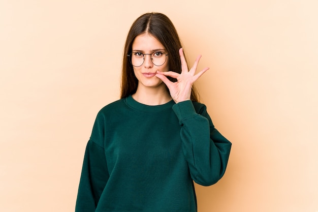 Jeune femme caucasienne sur mur beige avec les doigts sur les lèvres gardant un secret.